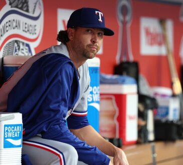 Texas Rangers pitcher Jacob deGrom sits on the dugout rail in a warmup jacket before a game.