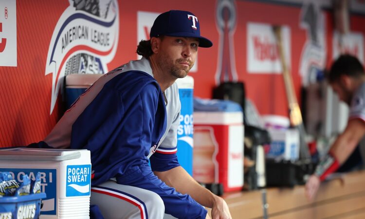 Texas Rangers pitcher Jacob deGrom sits on the dugout rail in a warmup jacket before a game.