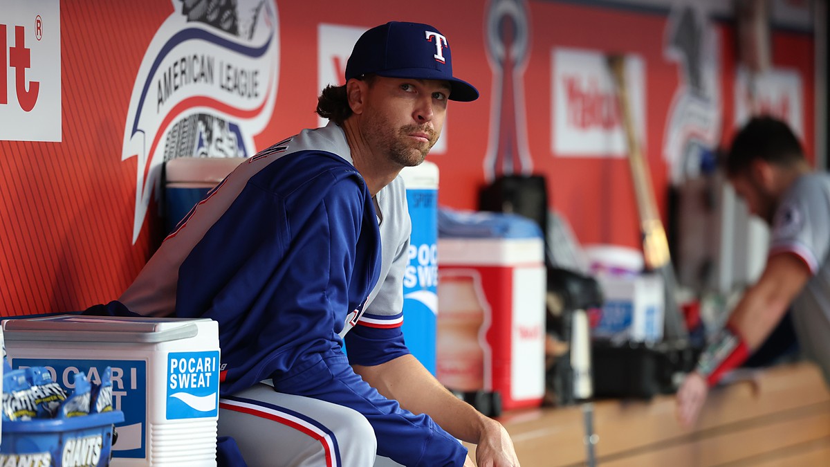 Texas Rangers pitcher Jacob deGrom sits on the dugout rail in a warmup jacket before a game.