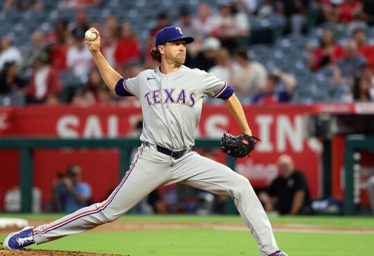 Texas Rangers pitcher Jacob deGrom strides toward the plate in road grays, ball hidden behind his hip.