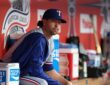 Texas Rangers pitcher Jacob deGrom sits on the dugout rail in a warmup jacket before a game.