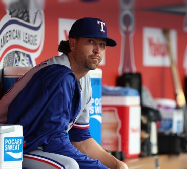 Texas Rangers pitcher Jacob deGrom sits on the dugout rail in a warmup jacket before a game.