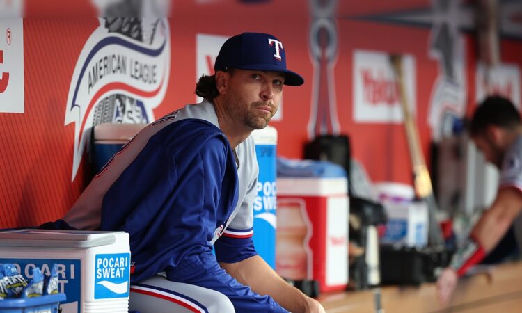 Texas Rangers pitcher Jacob deGrom sits on the dugout rail in a warmup jacket before a game.