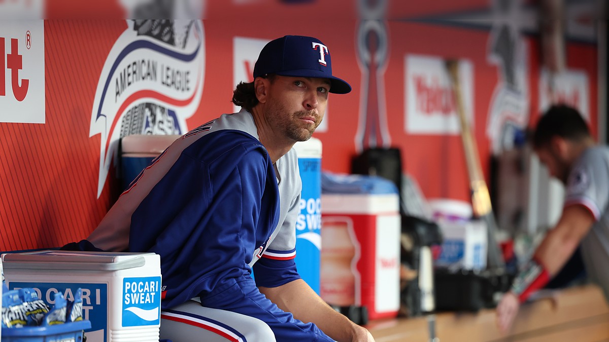 Texas Rangers pitcher Jacob deGrom sits on the dugout rail in a warmup jacket before a game.
