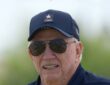 Dallas Cowboys owner Jerry Jones smiles in a team cap and aviator sunglasses at training camp.