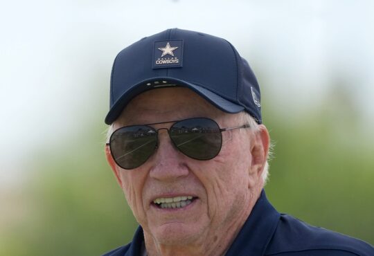 Dallas Cowboys owner Jerry Jones smiles in a team cap and aviator sunglasses at training camp.