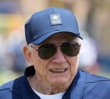 Dallas Cowboys owner Jerry Jones smiles on the practice field in a team cap and sunglasses at training camp.