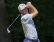 Golfer Jordan Spieth holds his follow-through with an iron after hitting a tee shot in a white cap and polo.