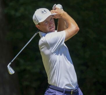 Golfer Jordan Spieth holds his follow-through with an iron after hitting a tee shot in a white cap and polo.