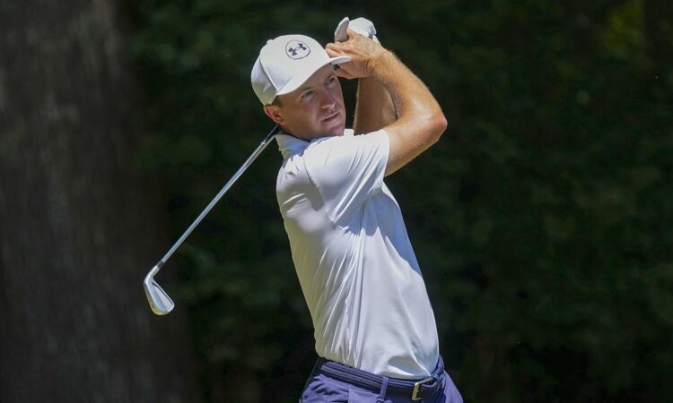 Golfer Jordan Spieth holds his follow-through with an iron after hitting a tee shot in a white cap and polo.