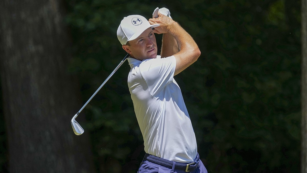 Golfer Jordan Spieth holds his follow-through with an iron after hitting a tee shot in a white cap and polo.