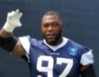 Dallas Cowboys defensive tackle Kenny Clark raises a taped hand while talking during training camp in a navy No. 97 jersey.
