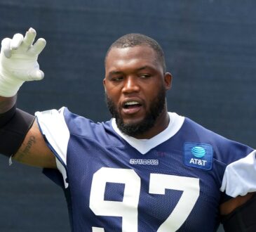 Dallas Cowboys defensive tackle Kenny Clark raises a taped hand while talking during training camp in a navy No. 97 jersey.