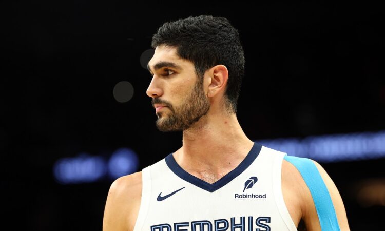 Memphis Grizzlies forward Santi Aldama looks toward the court in a white Memphis jersey with blue kinesiology tape on his shoulder.
