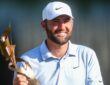 Scottie Scheffler smiles as he holds the FedEx St. Jude Championship trophy in a white cap and light blue polo.