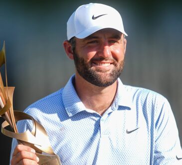 Scottie Scheffler smiles as he holds the FedEx St. Jude Championship trophy in a white cap and light blue polo.