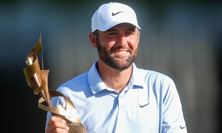Scottie Scheffler smiles as he holds the FedEx St. Jude Championship trophy in a white cap and light blue polo.