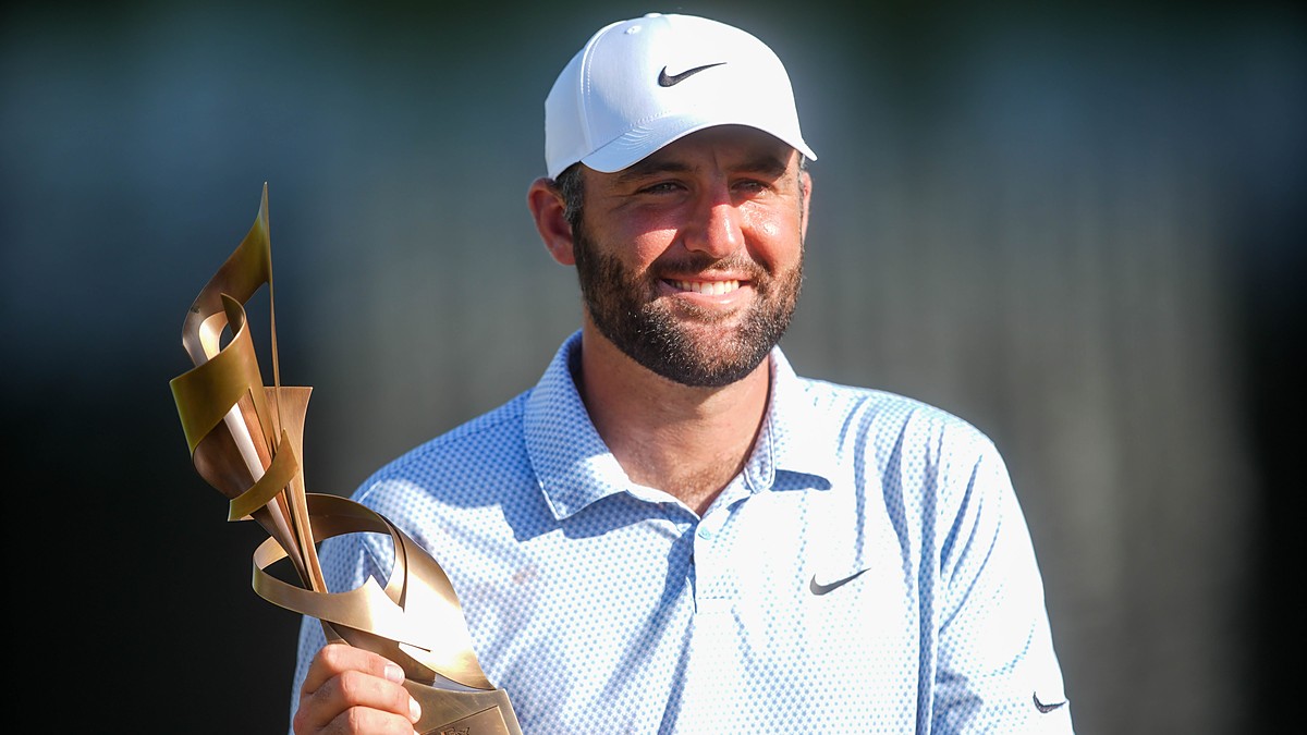 Scottie Scheffler smiles as he holds the FedEx St. Jude Championship trophy in a white cap and light blue polo.