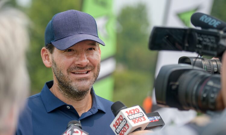 Former Dallas Cowboys quarterback Tony Romo smiles as he answers questions in front of television microphones.