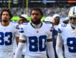 Dallas Cowboys wide receiver Traeshon Holden stands on the sideline in his white No. 80 jersey between two teammates.