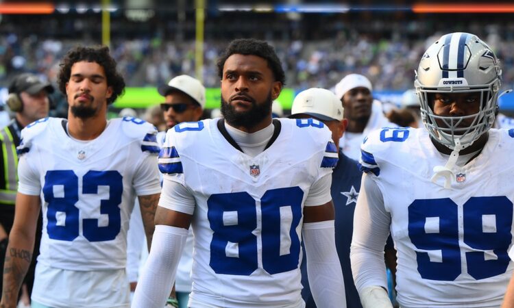 Dallas Cowboys wide receiver Traeshon Holden stands on the sideline in his white No. 80 jersey between two teammates.