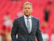 Pro Football Hall of Famer Troy Aikman stands on the field in a gray plaid suit before a Chiefs game.