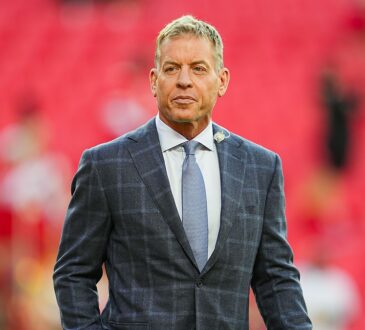 Pro Football Hall of Famer Troy Aikman stands on the field in a gray plaid suit before a Chiefs game.