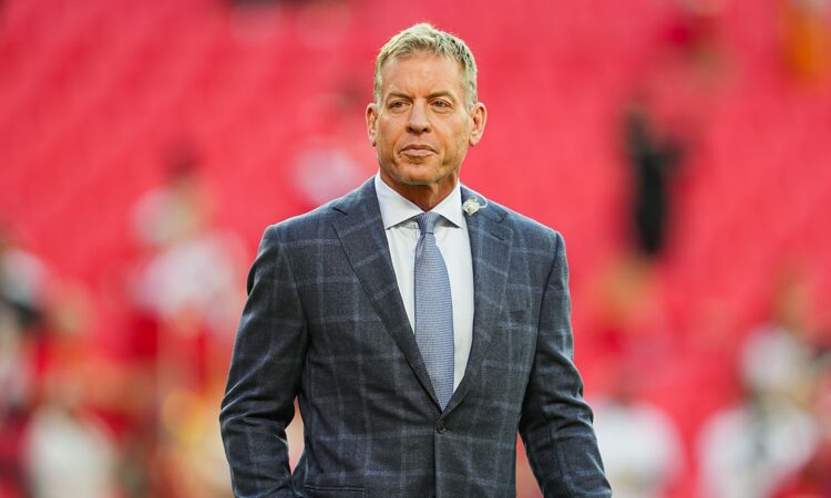 Pro Football Hall of Famer Troy Aikman stands on the field in a gray plaid suit before a Chiefs game.