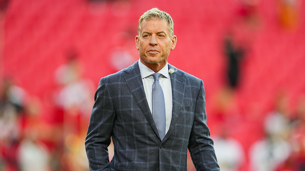 Pro Football Hall of Famer Troy Aikman stands on the field in a gray plaid suit before a Chiefs game.