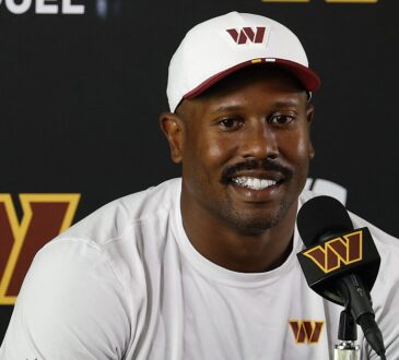 Washington Commanders outside linebacker Von Miller smiles while answering questions at a podium in a white team cap.
