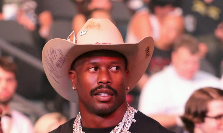 Von Miller looks on in a cream cowboy hat and diamond chains from the crowd at a UFC event.