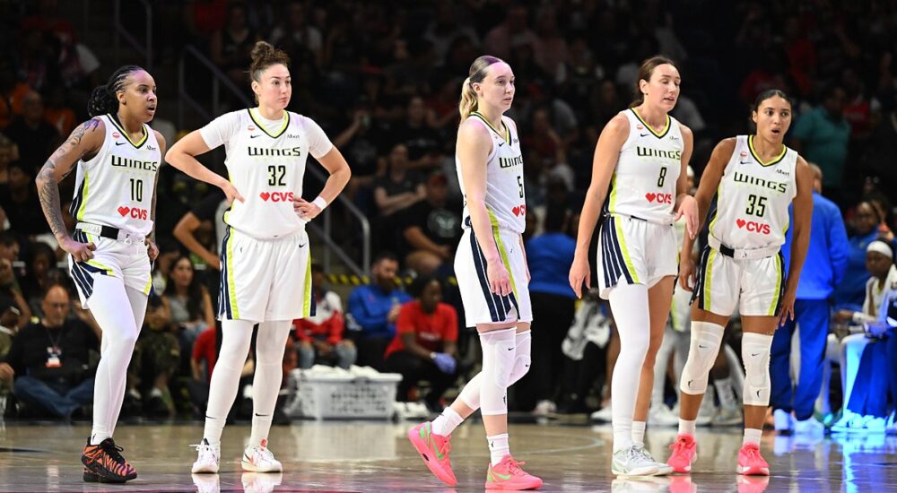 Dallas Wings guard Paige Bueckers and four teammates in white road uniforms stand on the court during a stoppage in play.