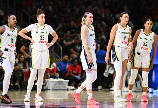 Dallas Wings guard Paige Bueckers and four teammates in white road uniforms stand on the court during a stoppage in play.