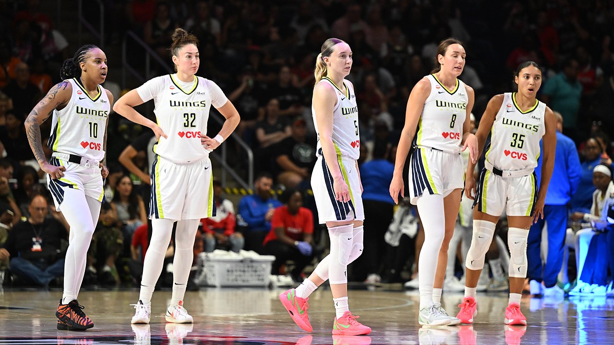 Dallas Wings guard Paige Bueckers and four teammates in white road uniforms stand on the court during a stoppage in play.