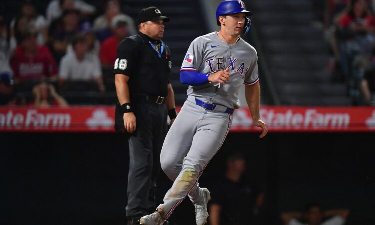 Texas Rangers left fielder Wyatt Langford jogs across the plate to score as the umpire watches behind him.