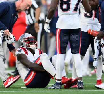 New England Patriots wide receiver A.J. Brown sits on the field as a trainer and teammates gather around him.