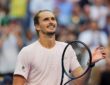 Tennis player Alexander Zverev smiles and raises a hand while holding his racket after winning a match.