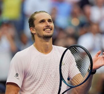 Tennis player Alexander Zverev smiles and raises a hand while holding his racket after winning a match.