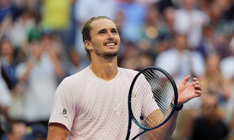 Tennis player Alexander Zverev smiles and raises a hand while holding his racket after winning a match.