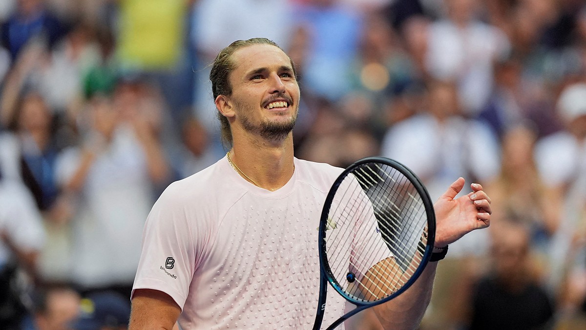 Tennis player Alexander Zverev smiles and raises a hand while holding his racket after winning a match.