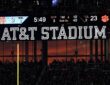 A sunset glows orange through the windows at AT&T Stadium beneath the scoreboard and the stadium name sign.