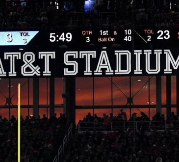 A sunset glows orange through the windows at AT&T Stadium beneath the scoreboard and the stadium name sign.