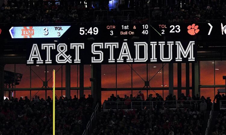 A sunset glows orange through the windows at AT&T Stadium beneath the scoreboard and the stadium name sign.