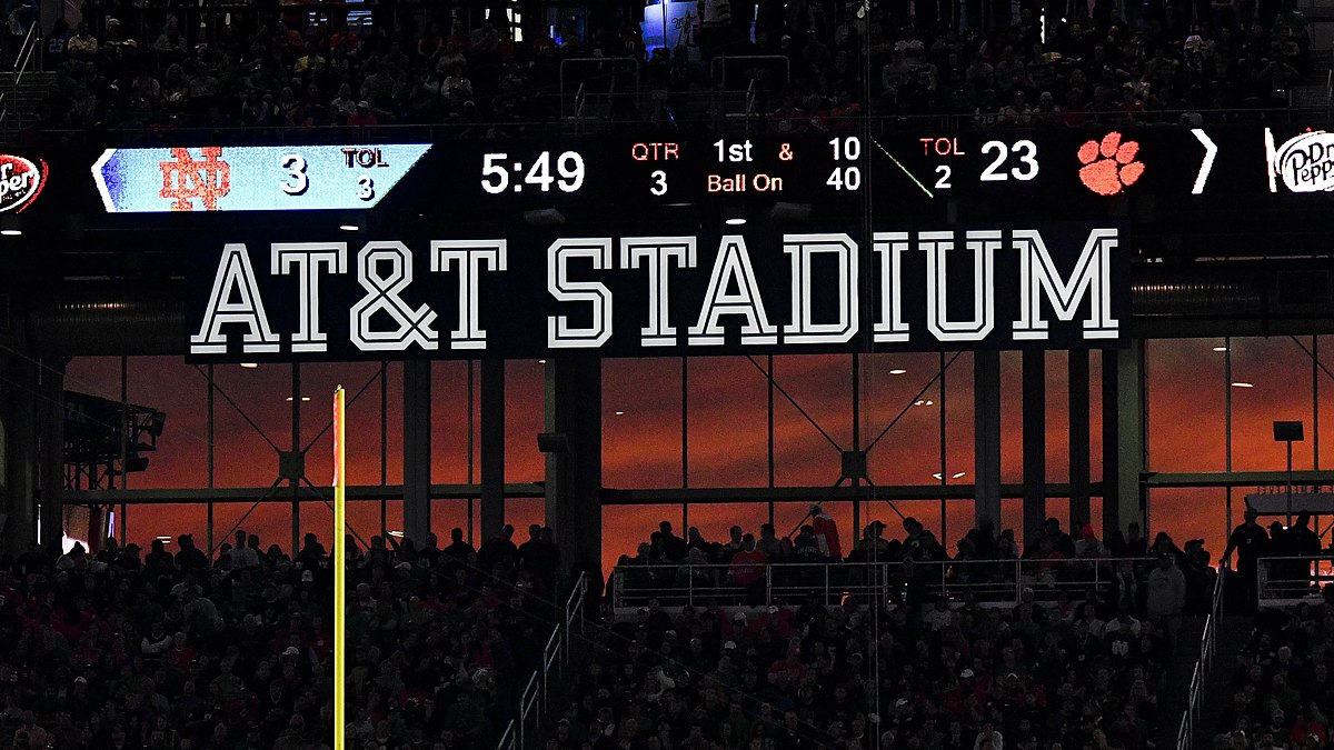 A sunset glows orange through the windows at AT&T Stadium beneath the scoreboard and the stadium name sign.