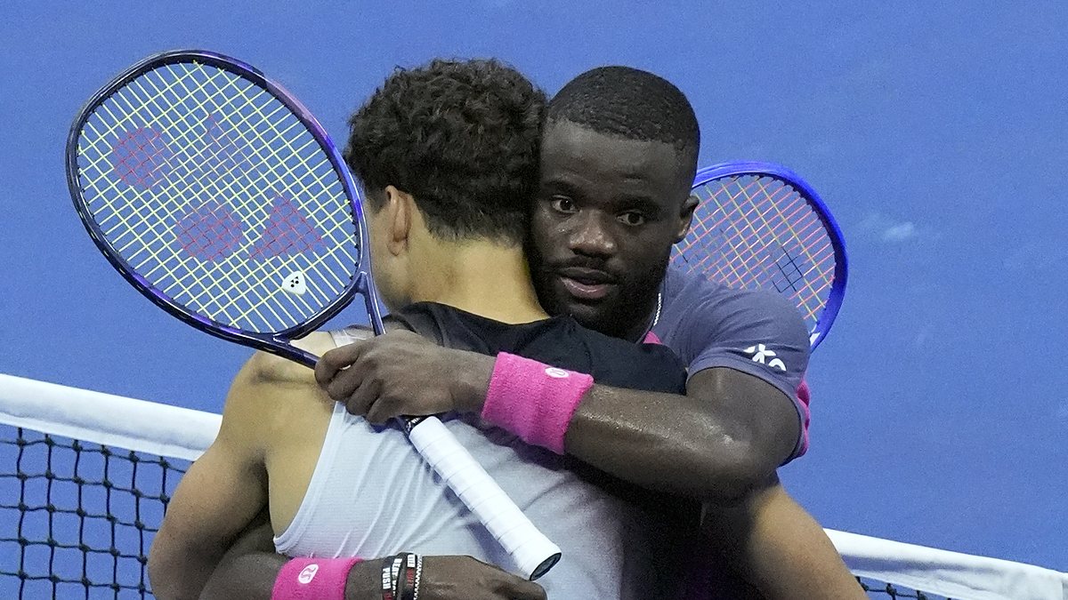 Ben Shelton, back to camera, and Frances Tiafoe hug at the net with their rackets raised after a match.