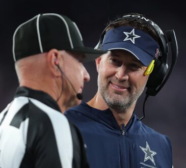 Dallas Cowboys head coach Brian Schottenheimer smiles while speaking with a game official on the sideline.