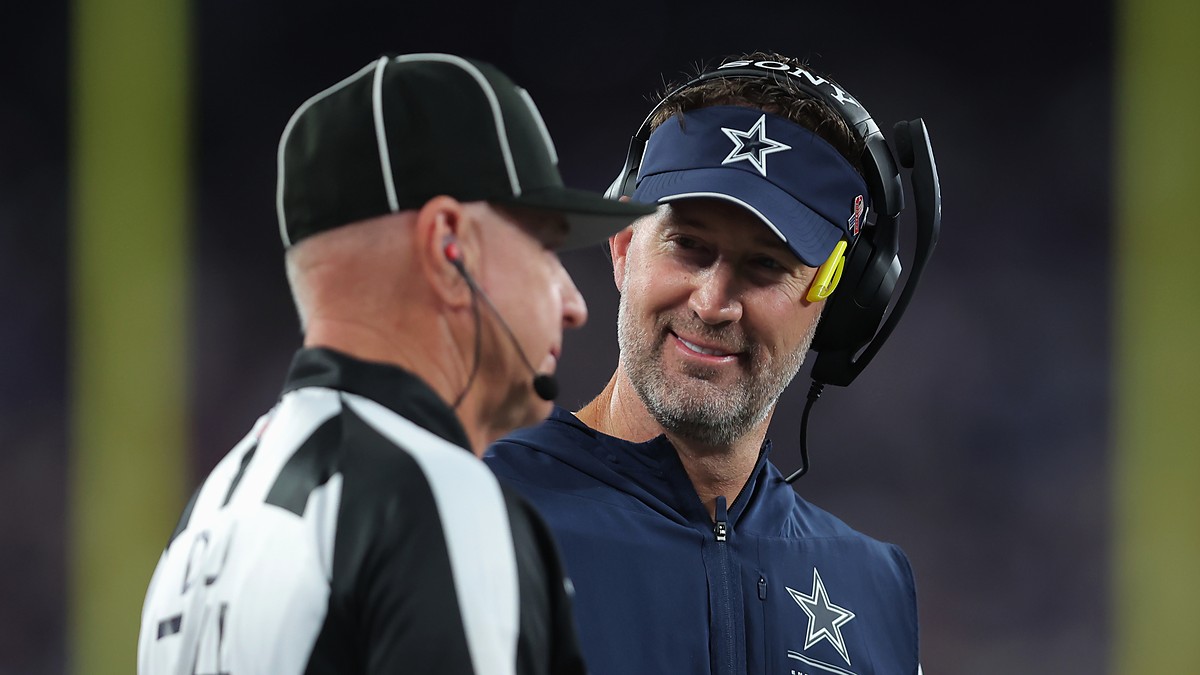 Dallas Cowboys head coach Brian Schottenheimer smiles while speaking with a game official on the sideline.