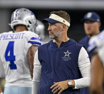 Cowboys head coach Brian Schottenheimer stands among players in warmups and looks toward the field before kickoff.