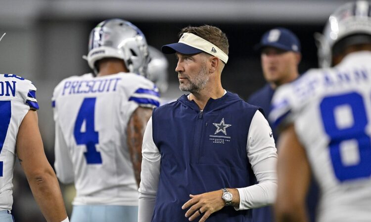 Cowboys head coach Brian Schottenheimer stands among players in warmups and looks toward the field before kickoff.
