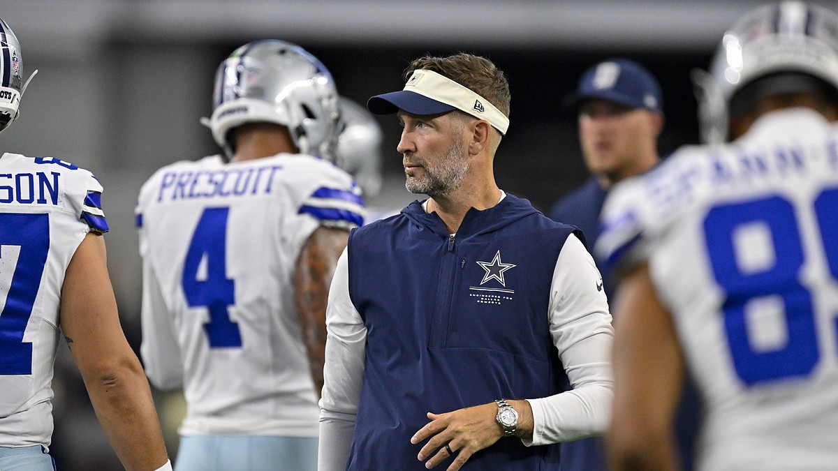 Cowboys head coach Brian Schottenheimer stands among players in warmups and looks toward the field before kickoff.